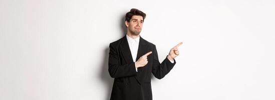 Image of upset and disappointed handsome guy in formal suit, pointing and looking left with sad face, standing over white background photo