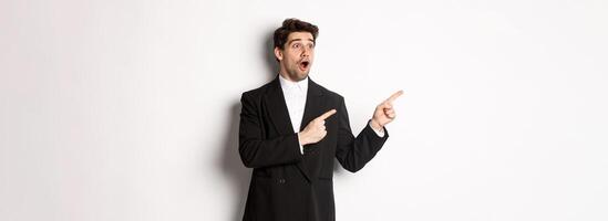 Impressed handsome man in party suit, looking at new year promo offer and pointing fingers left at banner, standing over white background photo