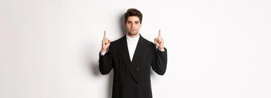 Image of confident and handsome man in formal suit, pointing fingers up, showing copy space on white background photo