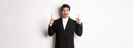 Image of handsome man in party suit, showing holidays promo, pointing fingers up and smiling amazed, standing over white background photo