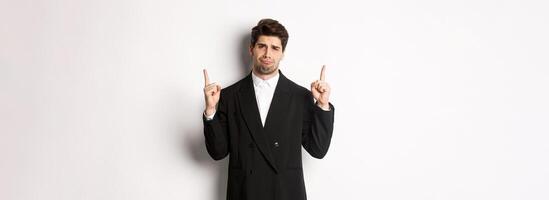 Portrait of reluctant and sad handsome man, complaining and pointing fingers up at something bad, looking jealous, standing over white background photo