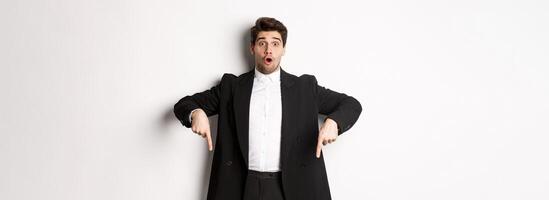 Portrait of surprised and excited man in black suit, pointing fingers down and showing christmas advertisement, standing over white background photo