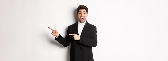Image of attractive smiling guy dressed for new year party, pointing fingers right and showing advertisement, looking impressed standing over white background photo