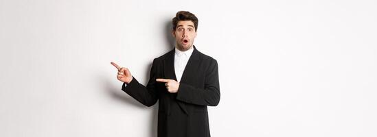 Portrait of surprised handsome man with beard, wearing black suit, open mouth and pointing fingers right at advertisement, standing over white background photo