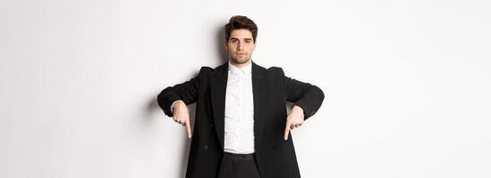 Image of handsome man dressed for formal party, wearing suit and pointing fingers down, showing advertisement or making announcement, standing over white background photo