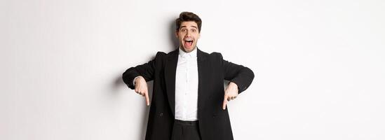 Portrait of super happy man showing christmas promo offer, pointing fingers down and smiling amazed, wearing formal party suit, standing over white background photo