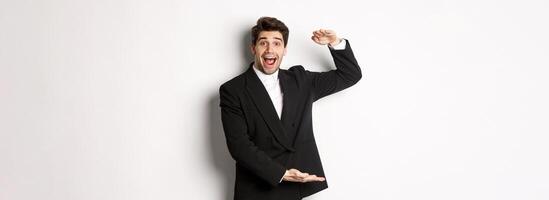 Portrait of excited handsome man in suit, shaping big object on copy space and smiling amazed, holding something, standing over white background photo
