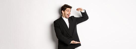 Portrait of excited handsome man in suit, shaping big object on copy space and smiling amazed, holding something, standing over white background photo