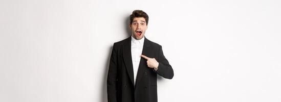Concept of new year party, celebration and lifestyle. Portrait of surprised handsome guy in black suit, pointing at himself and looking amazed, being chosen, standing over white background photo