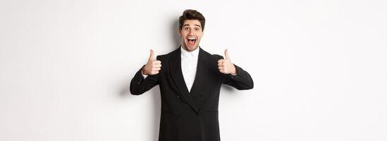 Concept of new year party, celebration and lifestyle. Portrait of amazed and pleased handsome man in black suit, showing thumbs-up, like product, approve something good, white background photo