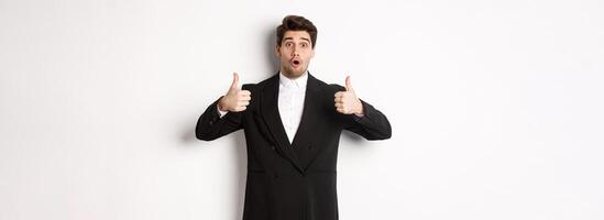 Concept of new year party, celebration and lifestyle. Portrait of excited handsome businessman in formal suit, showing thumbs-up in approval, recommending shop, standing over white background photo