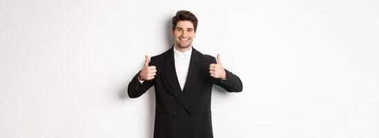 Concept of new year party, celebration and lifestyle. Image of attractive happy businessman in formal suit, showing thumbs-up and smiling, like and approve, standing over white background photo