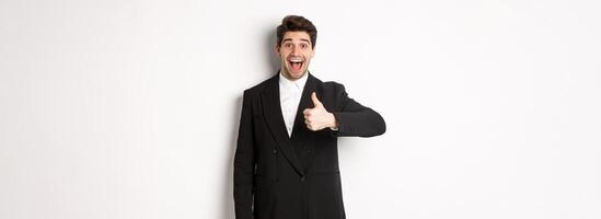 Portrait of amazed and impressed handsome man in black suit, showing thumb-up, praise awesome product, liking something good, standing against white background photo