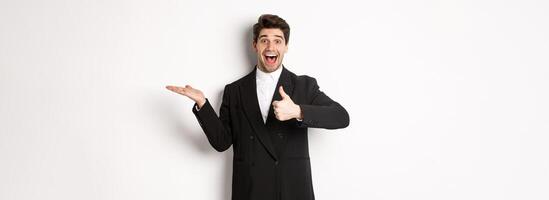 Portrait of hadnsome bearded man in formal suit, showing thumb-up and holding product in hand over white copy space, recommending product, standing over white background photo