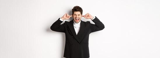 Portrait of annoyed and bothered businessman in black suit, shut ears and yelling, complaining loud noise, standing against white background photo