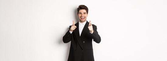 Concept of new year party, celebration and lifestyle. Handsome and successful guy in black suit, pointing fingers at camera and congratulating you, standing over white background photo