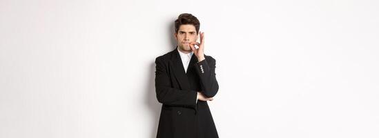Image of serious businessman in black suit, zipping mouth and frowning, hiding secret, showing taboo sign, standing over white background photo
