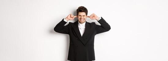 Portrait of annoyed and bothered businessman in black suit, shut ears, feeling uncomfortable, complaining loud noise, standing against white background photo