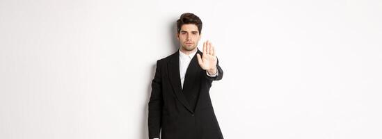 Portrait of serious handsome man in formal suit, extending hand to stop you, prohibit action, forbid and disagree with something, standing against white background photo