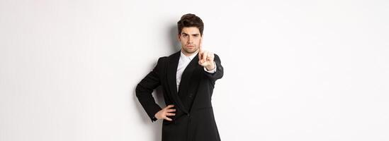 Portrait of serious handsome man in business suit, showing one finger to prohibit or decline something, telling to stop, disagree with you, standing over white background photo