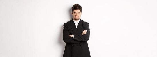Portrait of angry handsome man in black suit, cross arms on chest and looking offended, frowning and pouting, being mad at someone, standing over white background photo