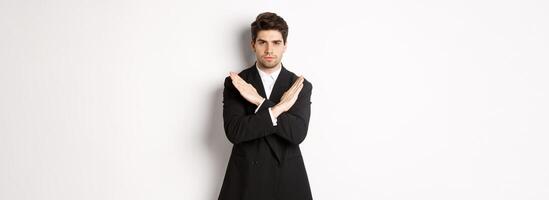 Portrait of serious and concerned man in black suit, showing stop gesture and frowning, making cross to prohibit or decline something, standing over white background photo