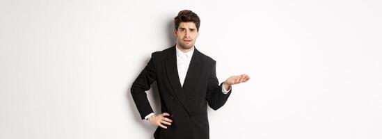 Image of confused handsome man in formal suit, raising hand and shrugging, cant understand something, standing against white background photo