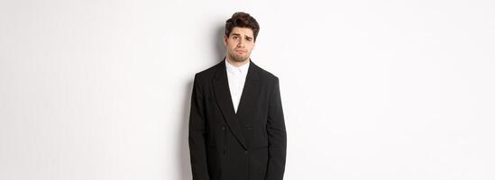 Image of gloomy and sad handsome male model in black suit, frowning and looking tired at camera, standing against white background photo