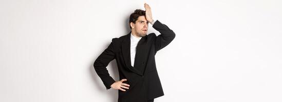 Portrait of shocked and distressed businessman in black suit, slap forehead and looking anxious, standing troubled over white background photo