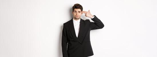 Portrait of distressed and troubled businessman in suit, pointing finger gun at head and looking bothered, shooting himself, standing against white background photo