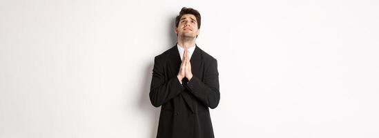Troubled and hopeful man in black suit begging god, pleading and looking up, need help, standing over white background photo