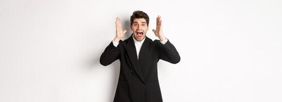 Frustrated and concerned man in black suit, screaming in panic and looking at something shocking, standing over white background photo