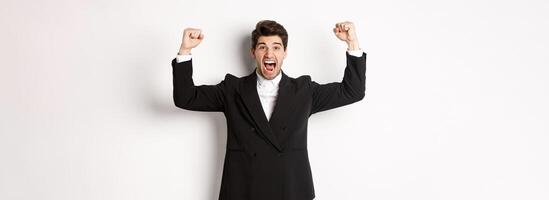 Successful handsome businessman triumphing, raising hands up and shouting yes, rejoicing over achievement, standing against white background photo