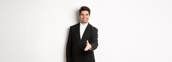 Portrait of handsome businessman in black suit, extending hand for handshake, greet business partners and smiling, welcome to company, standing over white background photo