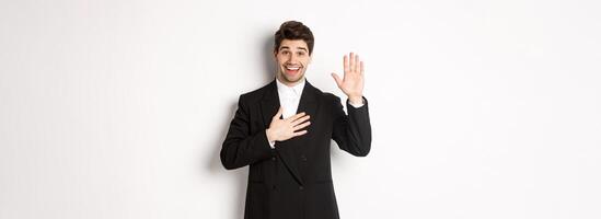 Image of handsome smiling guy telling the truth, raising on arm to promise something, standing over white background in black suit photo