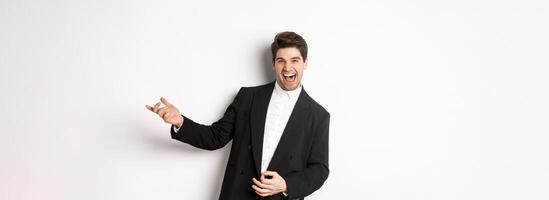 Portrait of happy man dancing at party, playing on invisible guitar and laughing, standing in black suit against white background photo