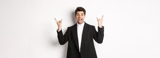 Portrait of happy attractive guy having fun at party, wearing black suit, showing rock-n-roll sign and tongue, standing excited against white background photo