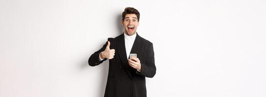 Portrait of handsome man in trendy suit showing thumbs-up in approval, using mobile phone app, smiling pleased, standing over white background photo