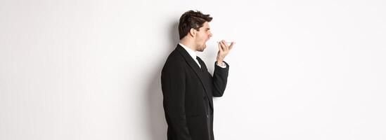 Profile shot of angry businessman in black suit, shouting at speakerphone and looking mad, recording voice message, standing over white background photo