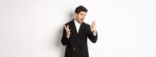 Portrait of angry businessman in black suit yelling at mobile phone, having an argument on call, standing mad over white background photo