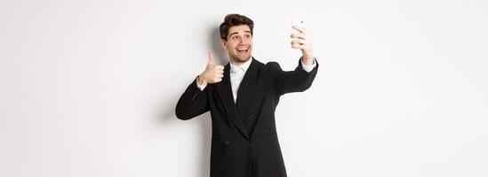 Portrait of good-looking man taking selfie on new year party, wearing suit, taking photo on smartphone and showing thumbs-up, standing against white background
