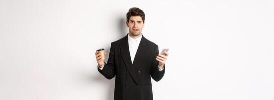 Image of confused businessman standing in a suit, holding cup of coffee and mobile phone, cant understand something, standing against white background photo