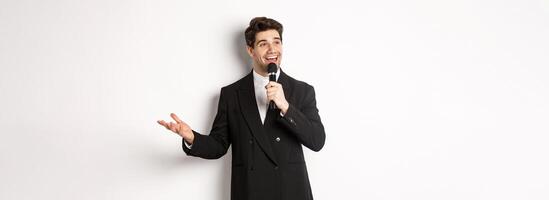 Portrait of handsome man in black suit singing a song, holding microphone and giving speech, standing against white background photo