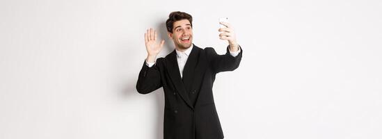 Image of handsome man in suit, having call and waving hand at smartphone camera, recording , greeting someone, standing against white background photo