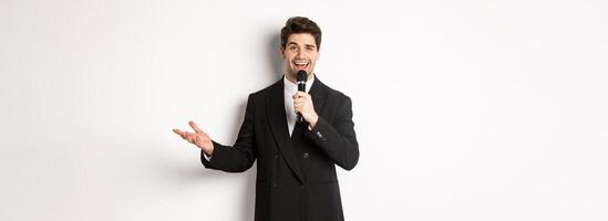 Portrait of handsome man in black suit singing a song, holding microphone and giving speech, standing against white background photo