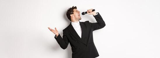 Portrait of handsome man singing a song with passion, standing in black suit, holding microphone and performning, posing over white background photo