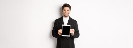Portrait of handsome businessman in trendy suit, showing digital tablet screen and smiling, standing against white background photo