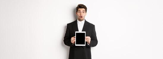 Image of surprised handsome man in black suit, showing digital tablet screen and looking amazed, standing against white background photo
