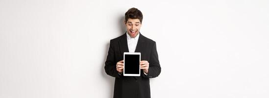 Portrait of amazed handsome man in black suit, showing digital tablet screen with excitement, standing over white background photo