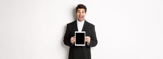 Image of attractive male entrepreneur in trendy suit, showing digital tablet screen and smiling amazed, standing over white background photo